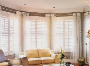 White shutters and sheer curtains in a bay window with a cream sofa